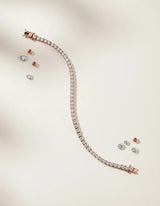 Pulsera Tennis Elaine Vault de Oro Rosa 14k, en toma de estudio, extendida en curva sobre fondo claro, con diamantes sueltos alrededor.