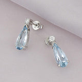 Aretes Aguamarina Corte Pera de Oro Blanco 14k, en toma de estudio sobre tela lila.
